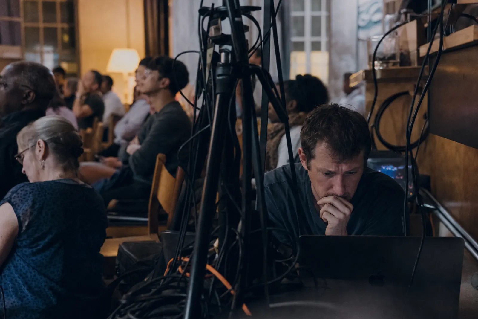 A stressed volunteer crouches behind tangled cables and a tripod, frowning at a laptop.