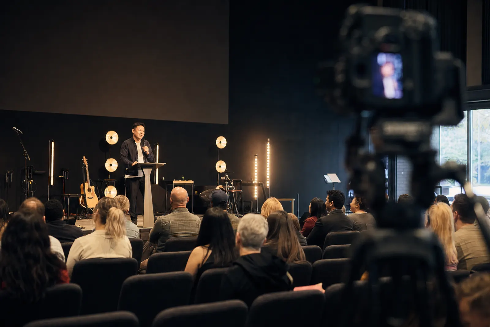 A camera at the back of the room frames the pastor preaching on a warmly lit stage.